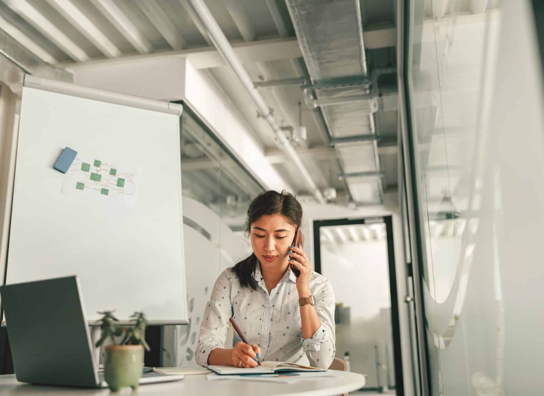 Woman office manager talking phone with client and making notes while sitting the desk in office